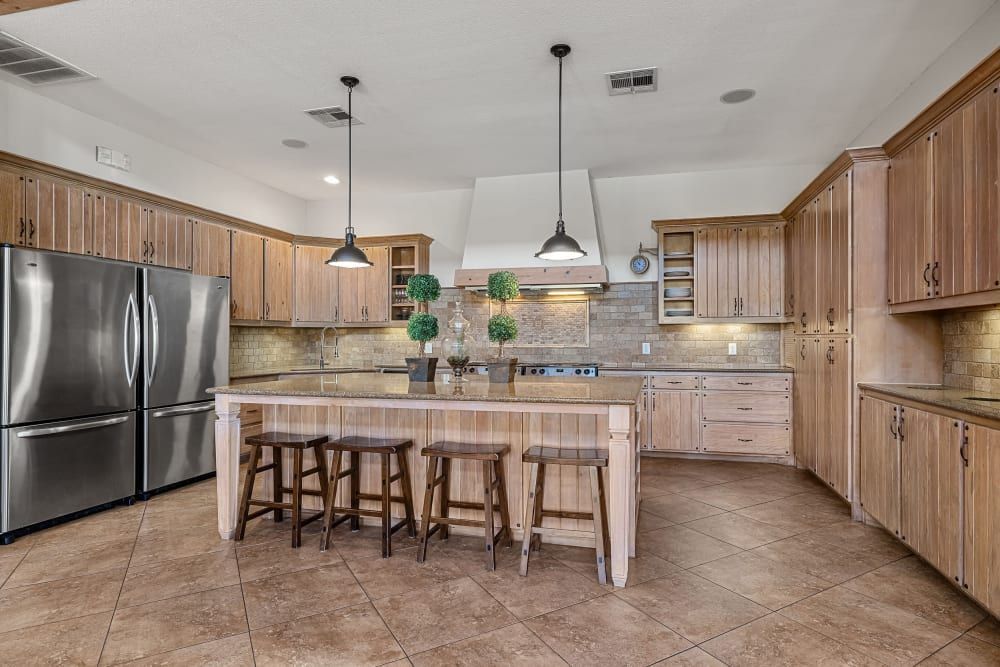 Spacious kitchen interior featuring wooden cabinets with black handles, stainless steel appliances, a central island with bar stools, and pendant lighting. The room has a beige backsplash, tiled flooring, and a bright atmosphere at Marquis at Canyon Ridge in Austin, TX.