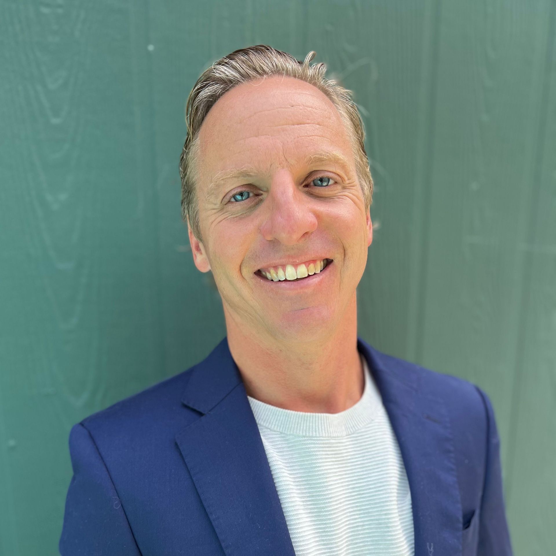 A smiling man with blond hair in a blue blazer and white shirt, standing against a teal-green background.
