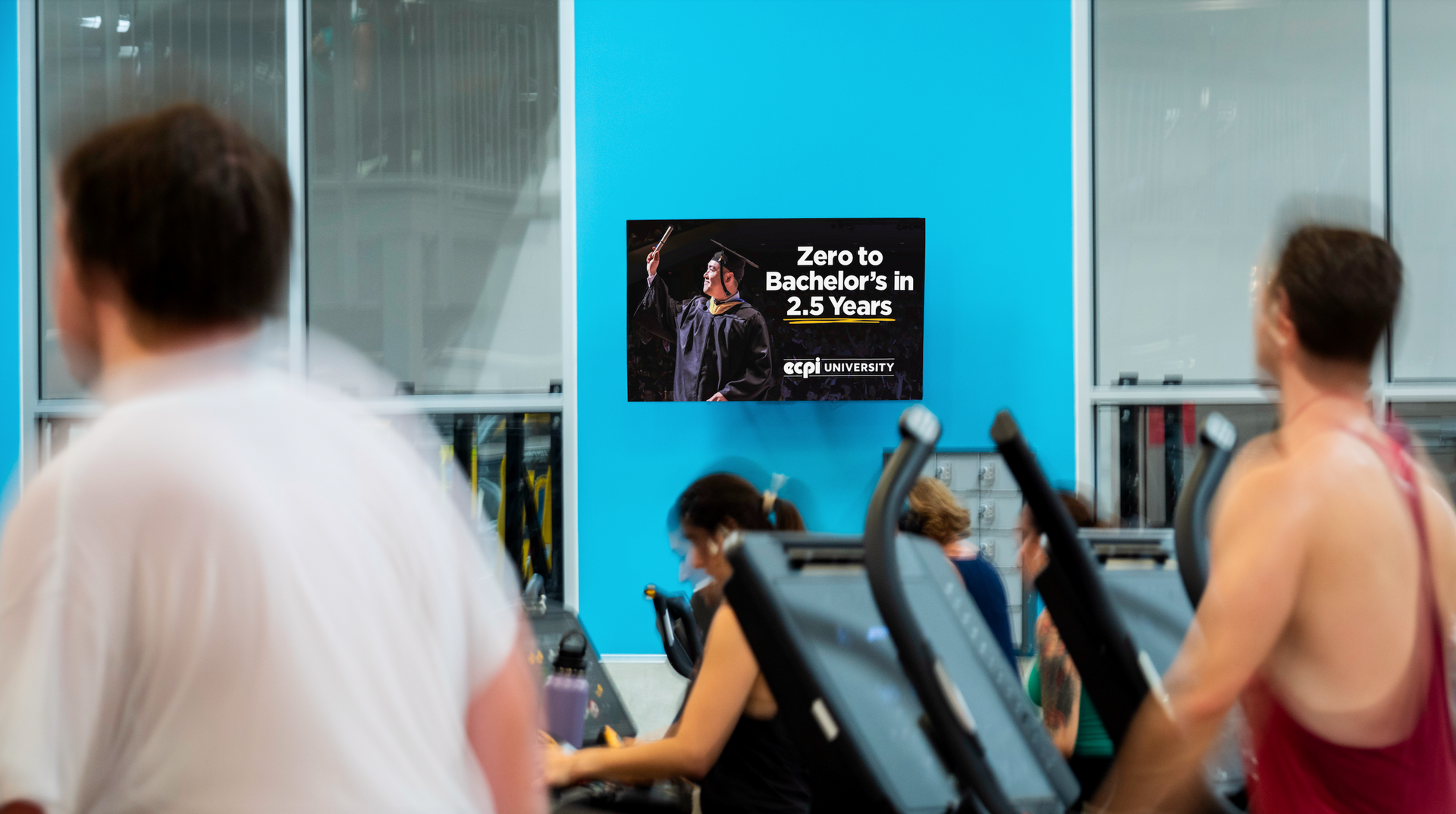 People on treadmills at a gym, looking at a TV screen on a blue wall. The screen displays an advertisement.
