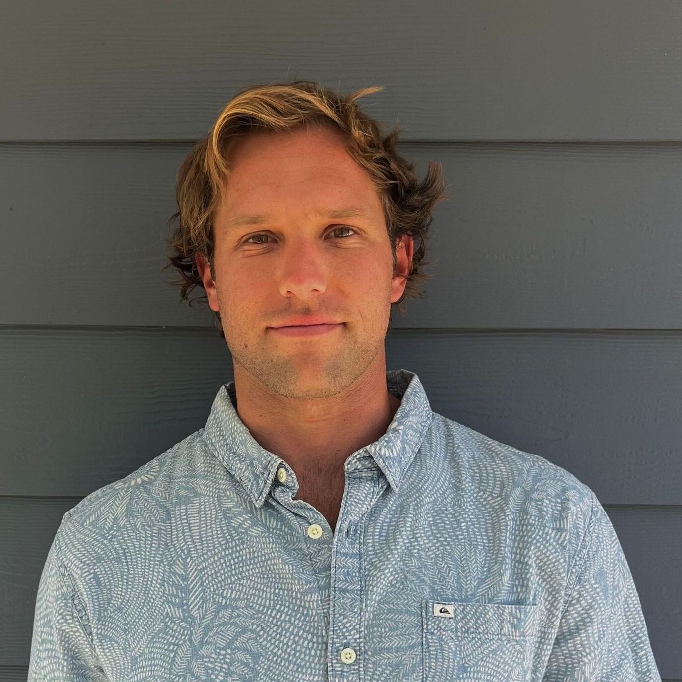 A man with wavy blonde hair wearing a blue patterned button-down shirt smiles in front of a blue wall.
