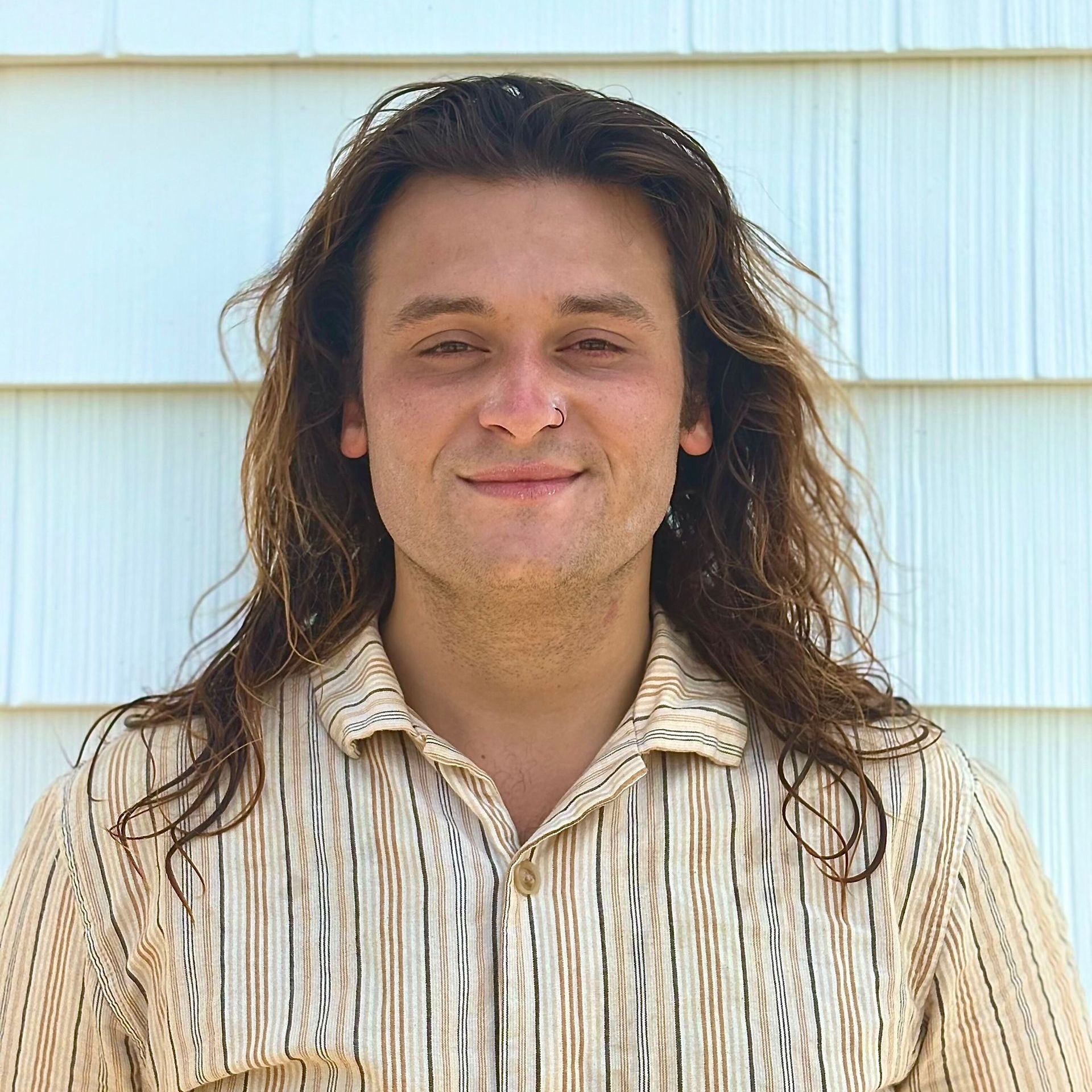 Man with long brown hair and nose ring, smiling in front of a white-sided building. He wears a tan striped shirt.