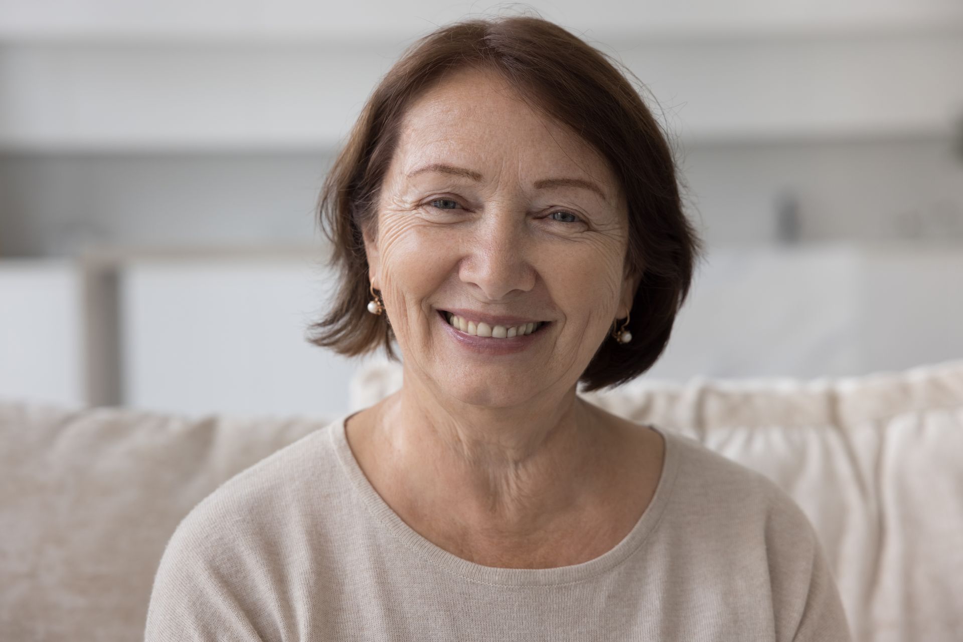An elderly woman is smiling while sitting on a couch.