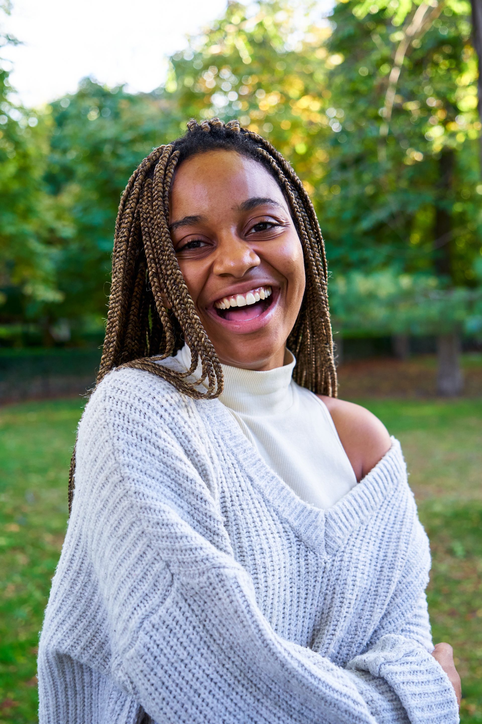 A woman with braids is wearing a sweater and smiling in a park.