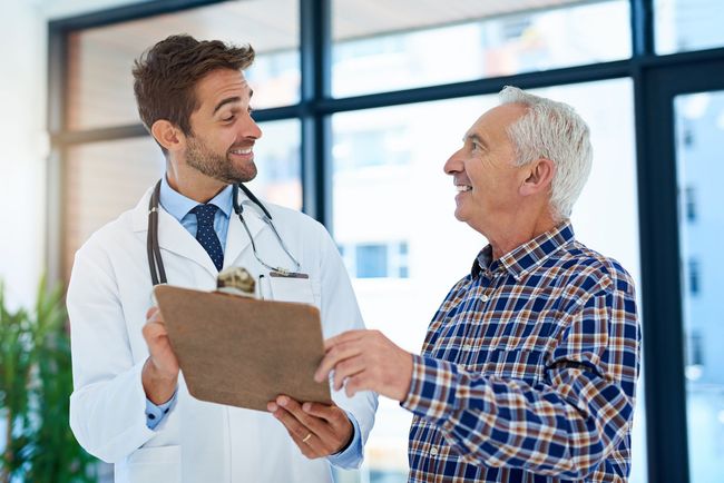 A doctor is talking to an older man while holding a clipboard.