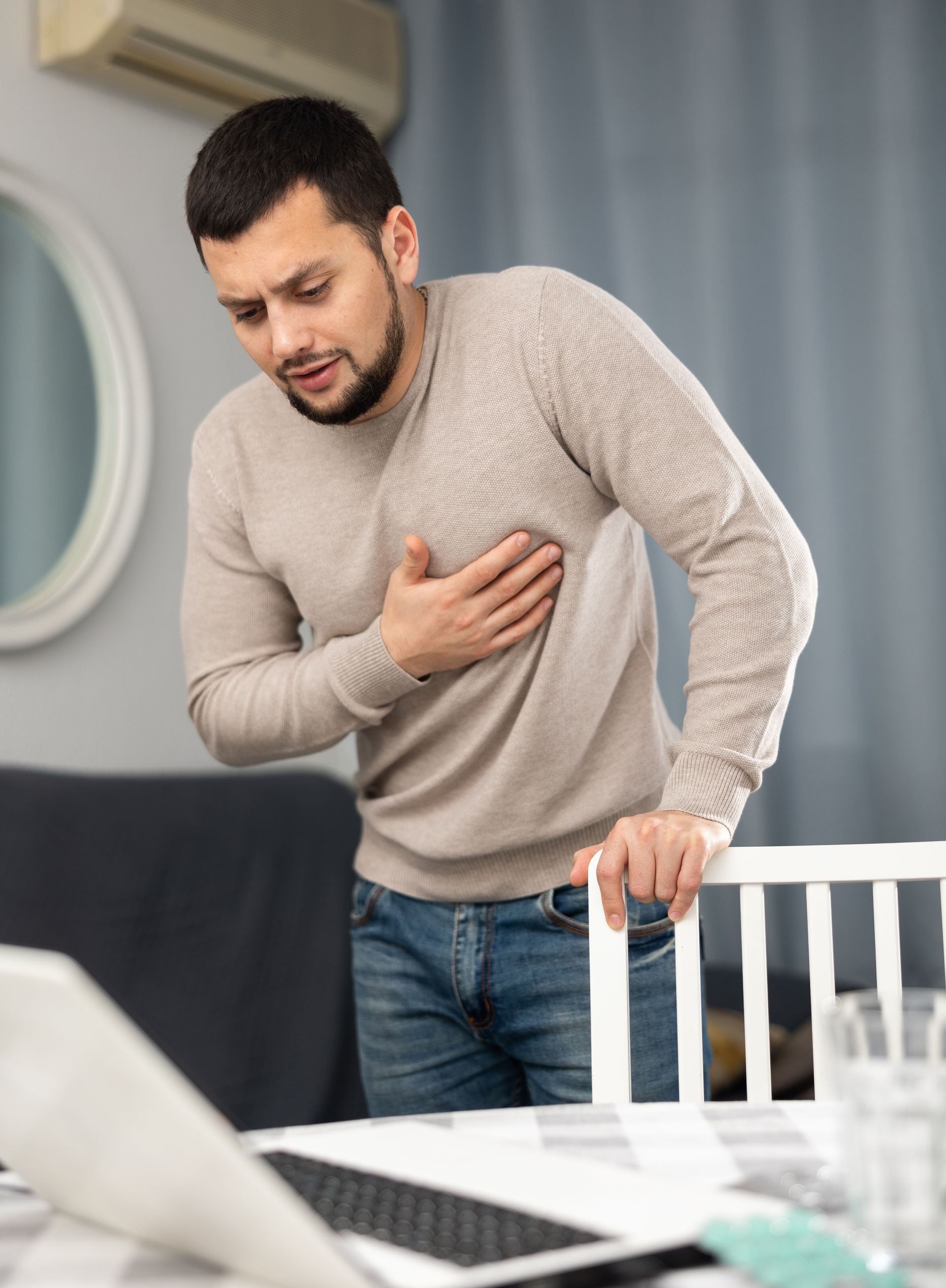 A man is holding his chest while standing in front of a laptop computer.