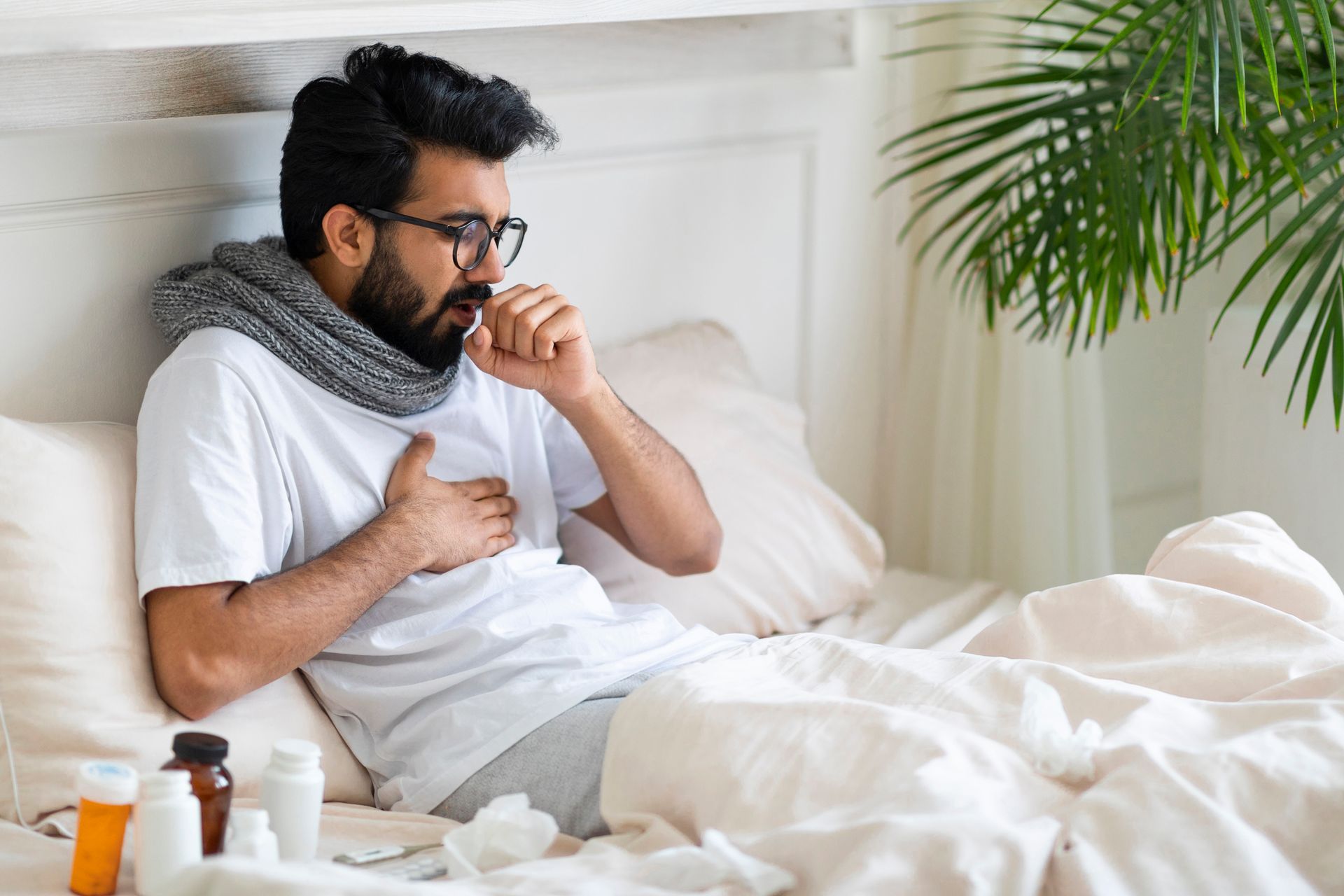 Man coughing in bed, wearing a scarf. Medications on bedside table.
