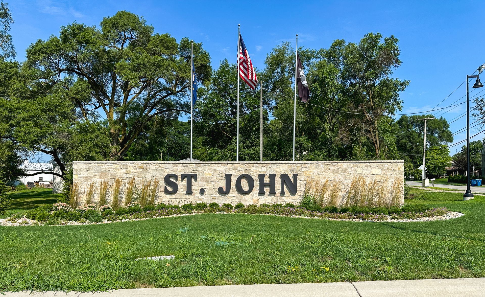Stone sign reading “ST. JOHN” with American flags and trees under a clear blue sky