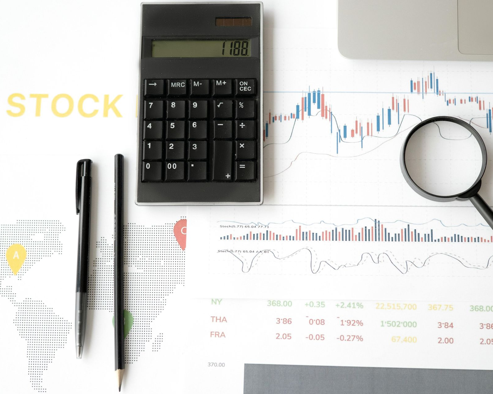Calculator, pens, and a magnifying glass resting on financial stock market charts and data tables on a white desk.