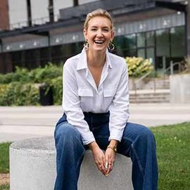 Woman in white shirt and jeans smiles, sitting on a concrete cylinder outdoors.