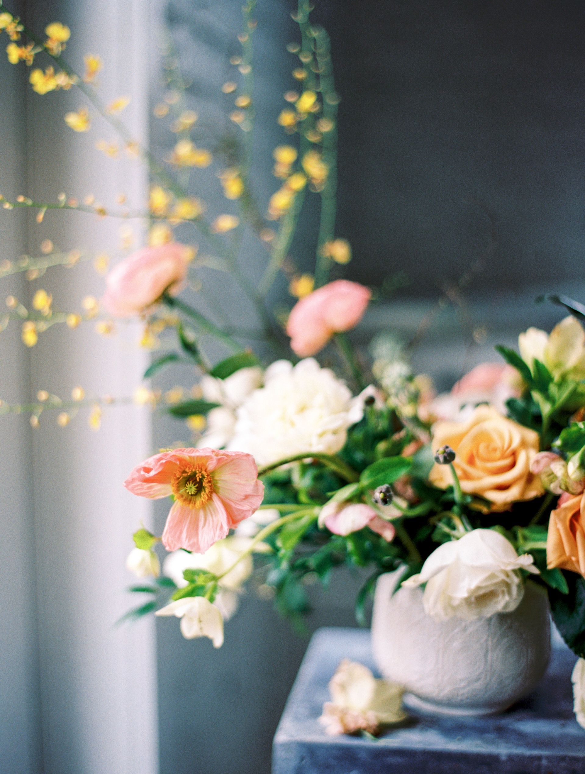 Close-up of floral arrangement in a stone vase: peach and white blooms with greenery, set against a muted background.
