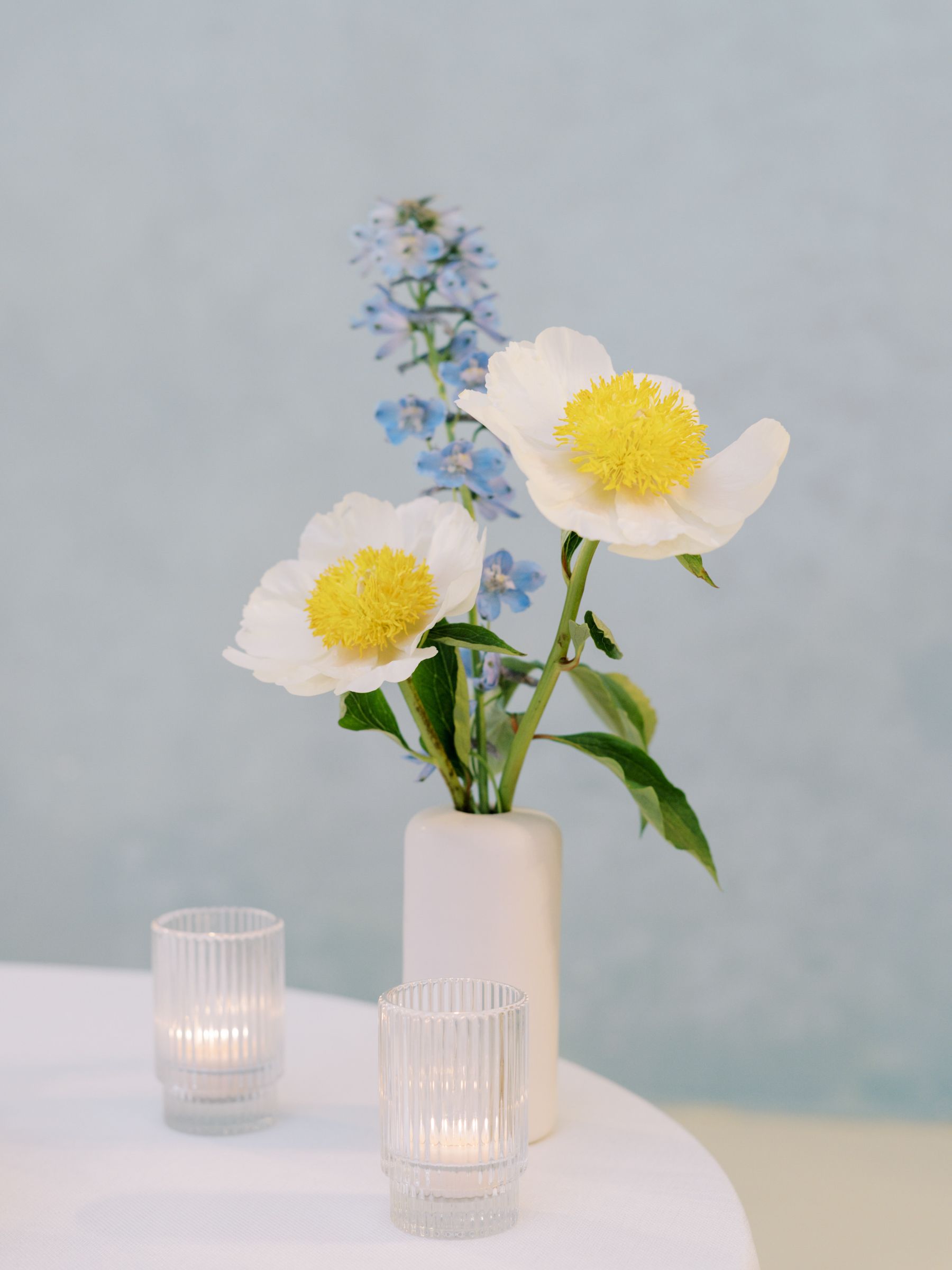 Showers - White flowers with yellow centers and blue flowers in a white vase on a white table with candles.