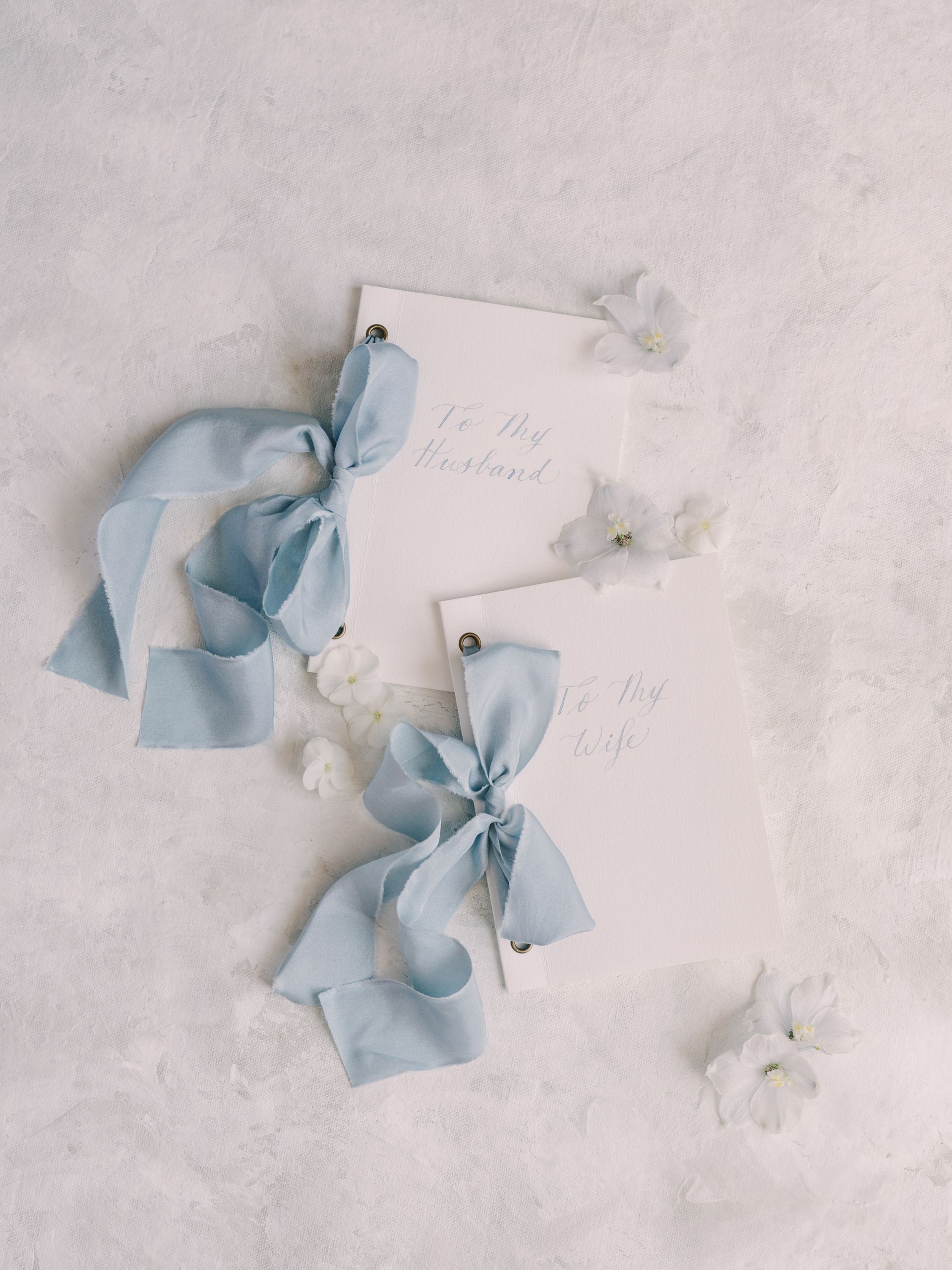Two white cards tied with blue ribbons, decorated with white flowers on a textured white background.