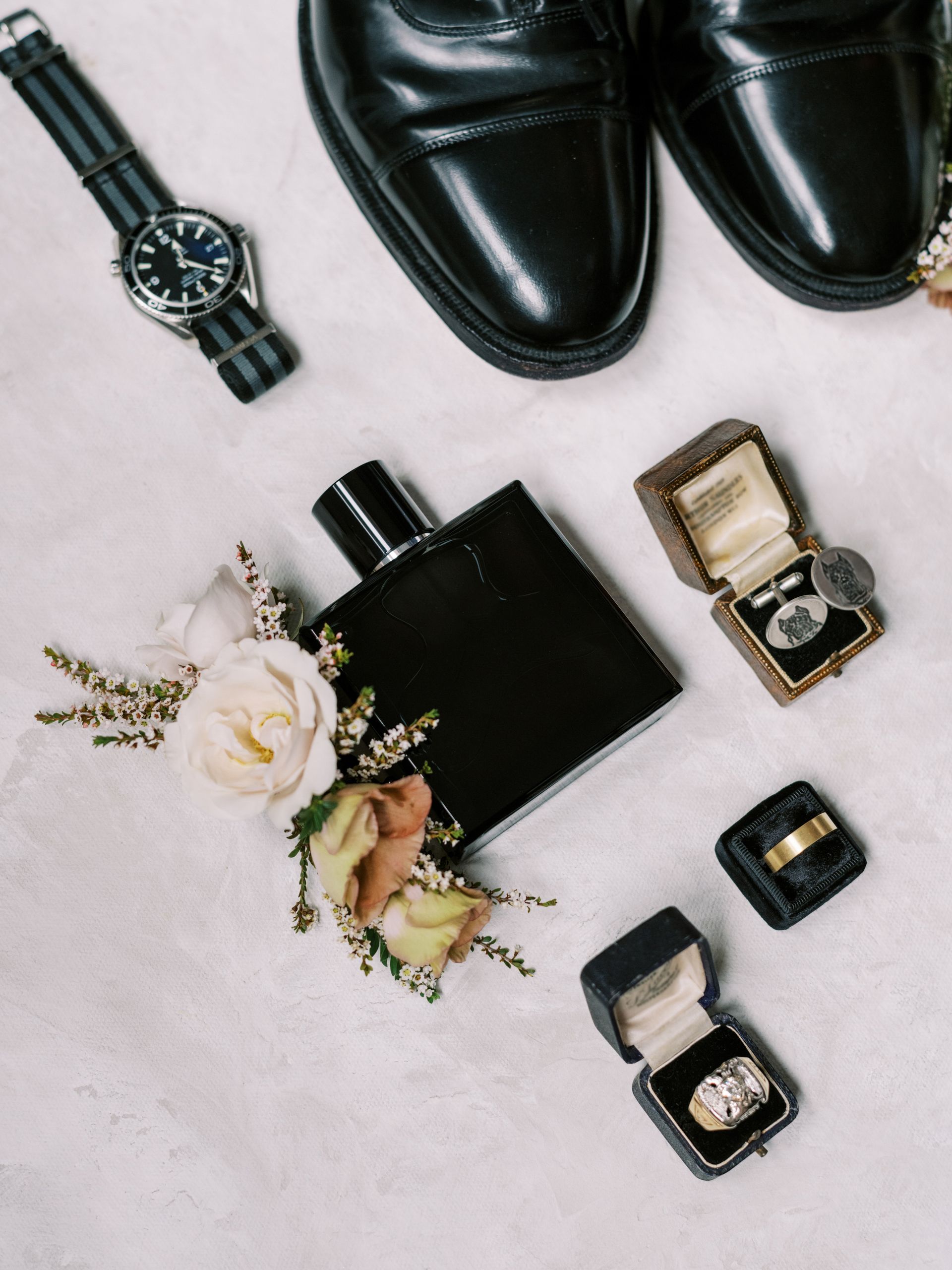 Men's accessories on white surface: black shoes, watch, cologne, rings, and floral arrangement.