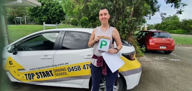 Person holding a P-plate with a car in front of a driving school car.  Setting is outdoors, daytime.