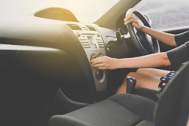 Person adjusting a car's climate control while driving. The interior is dark with sunlight streaming in.