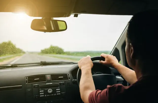 Driver holding steering wheel, driving on road, sunlight, interior car view.