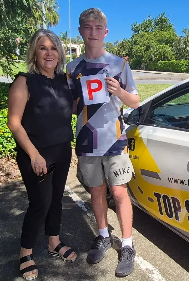 Teenage boy holds driving permit next to woman, standing by a car on a sunny day.