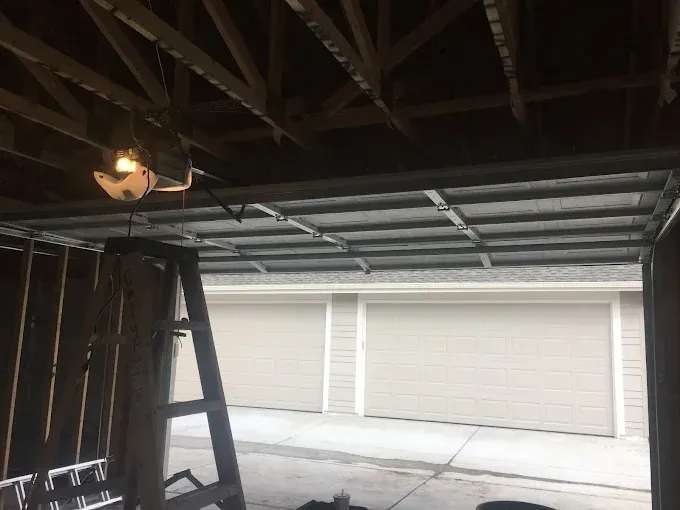 Garage interior with exposed ceiling beams, a ladder, and two white roll-up doors.
