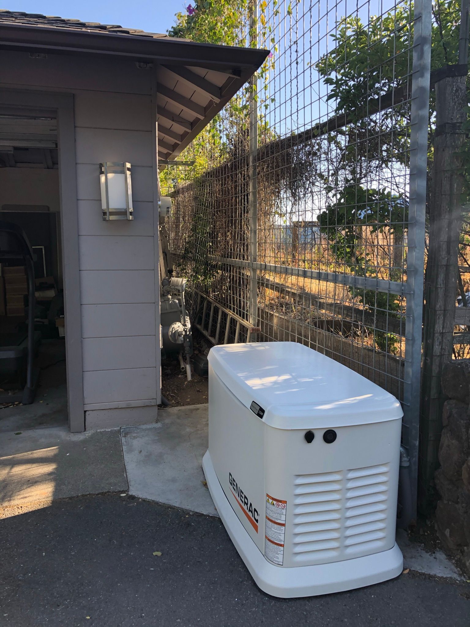 A generator is sitting outside of a garage next to a fence.