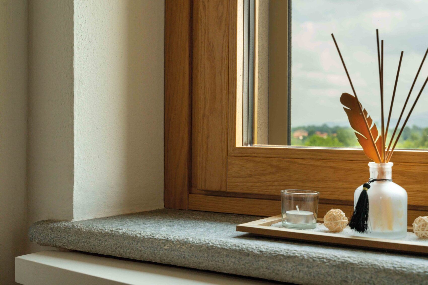 Un diffusore di aromi con bastoncini e una piccola candela sono posizionati su un davanzale di pietra accanto a una finestra con cornice in legno.
