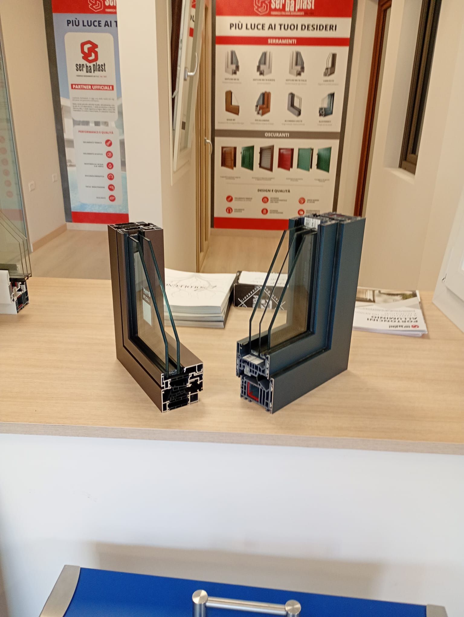 Due campioni di profili per finestre con cornice scura, che mostrano gli strati di vetro, sono appoggiati su un bancone di legno in uno showroom.