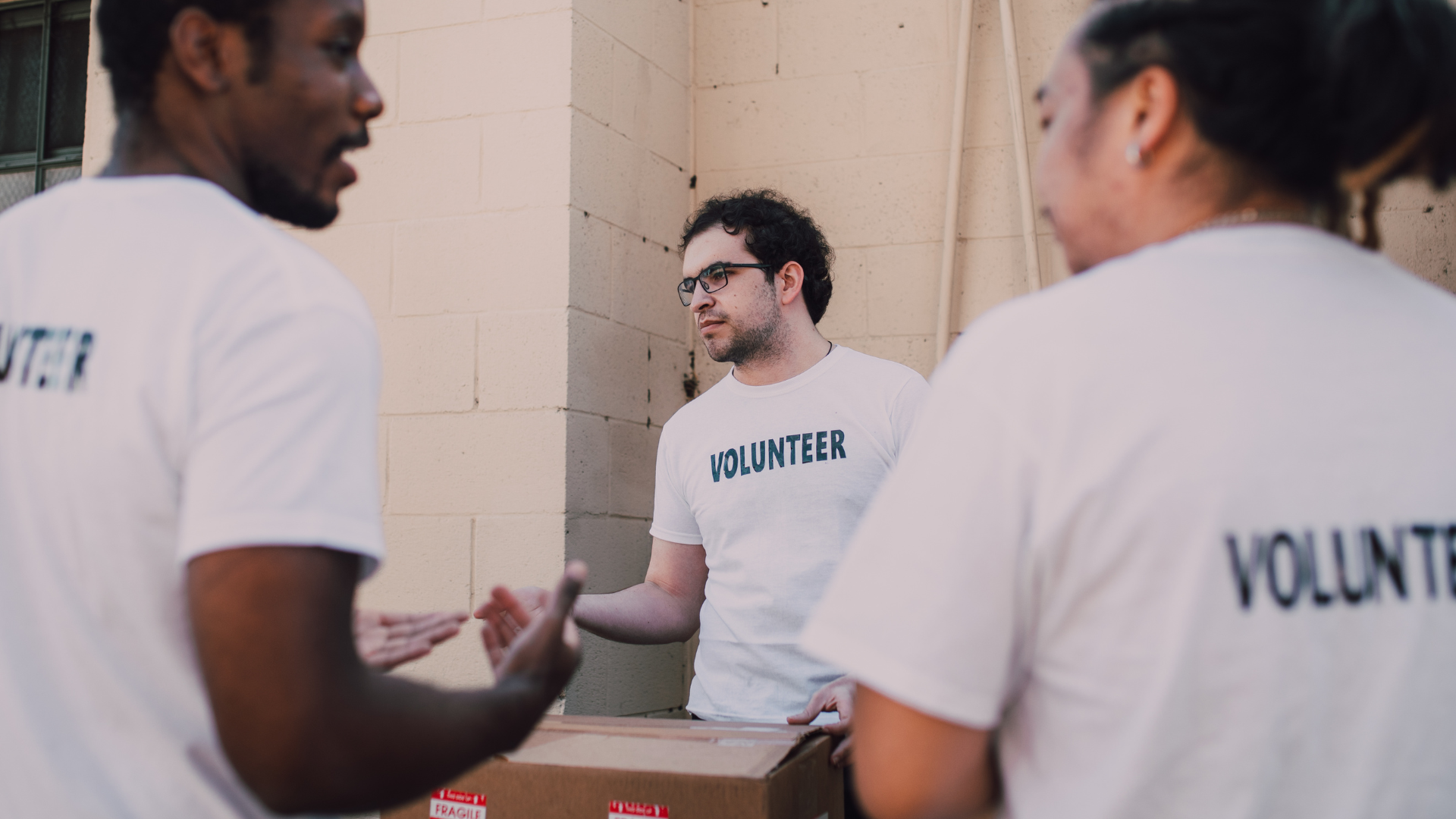 Volunteers carrying a cardboard box. One man gestures while another looks on, all wearing white 