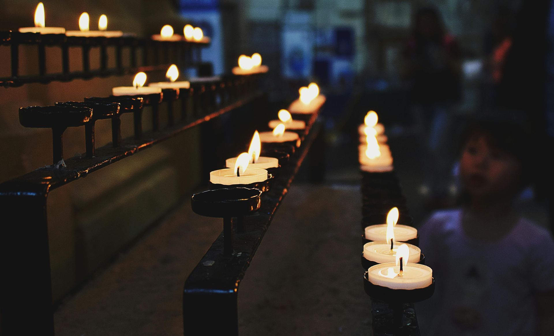 Candles lit in a dark church, burning on a black metal rack. A child is visible in the background.