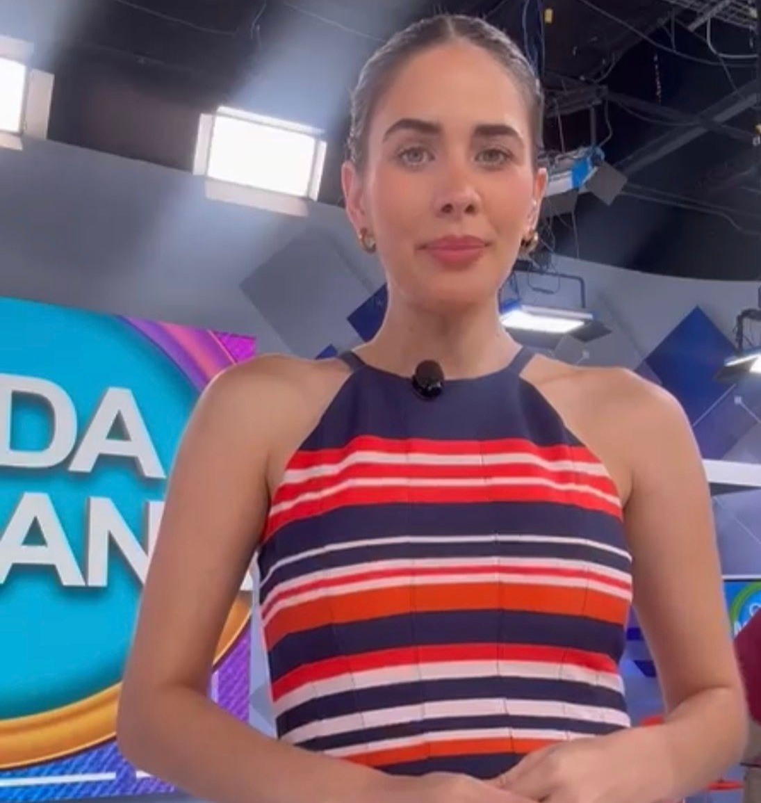 Presentadora de pie en un estudio de televisión, con un vestido de tirantes a rayas, mirando a la cámara.