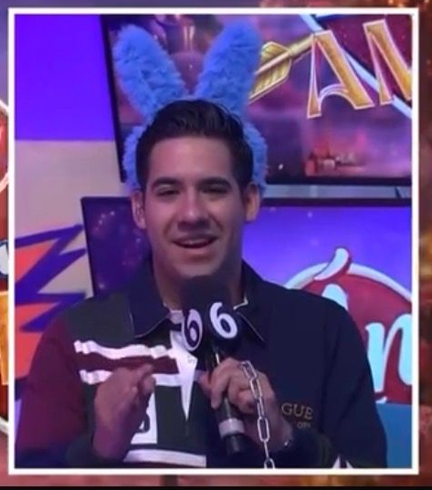 Un hombre con orejas de conejo azules, situado en un podio, habla por un micrófono en un colorido televisor.