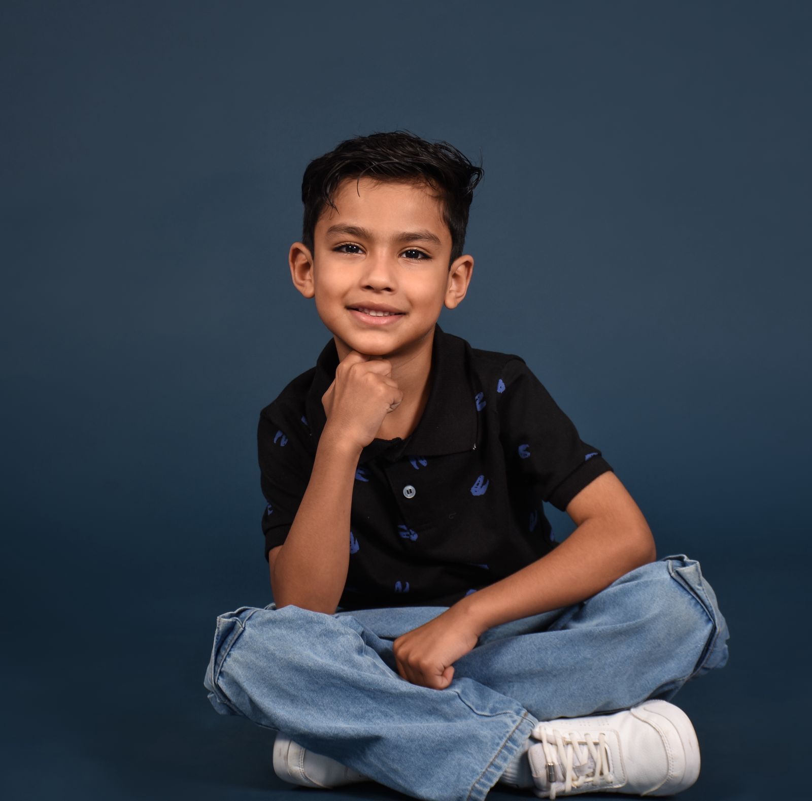 Niño sonriente sentado con las piernas cruzadas sobre un fondo azul, vistiendo una camiseta negra, vaqueros y zapatillas blancas.
