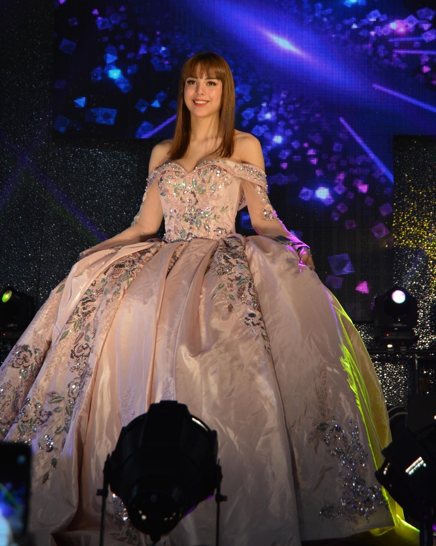Una mujer con un vestido brillante se encuentra en el escenario bajo luces violetas, extendiendo una falda de tul voluminosa.
