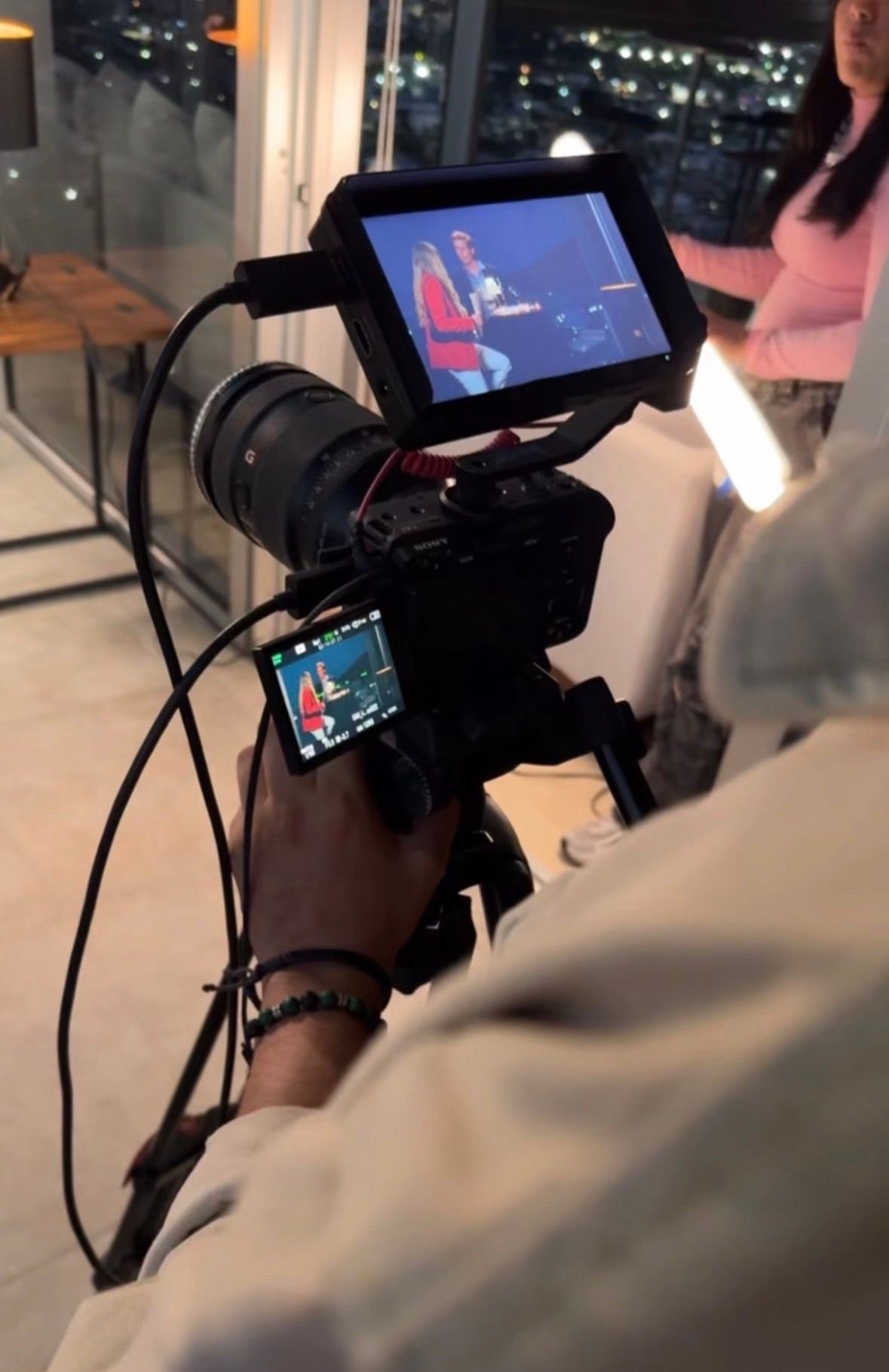 Una persona filma con un monitor de cámara en un interior con poca luz; las luces de la ciudad son visibles a través de las ventanas.