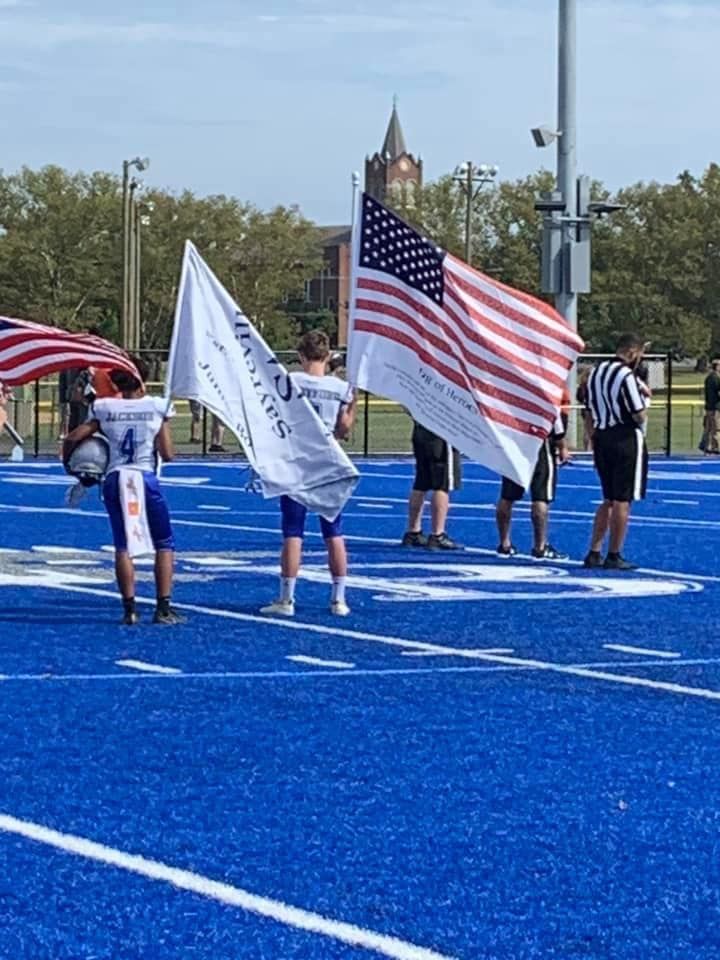 Football players hold flags on a blue field before a game. One flag is American.