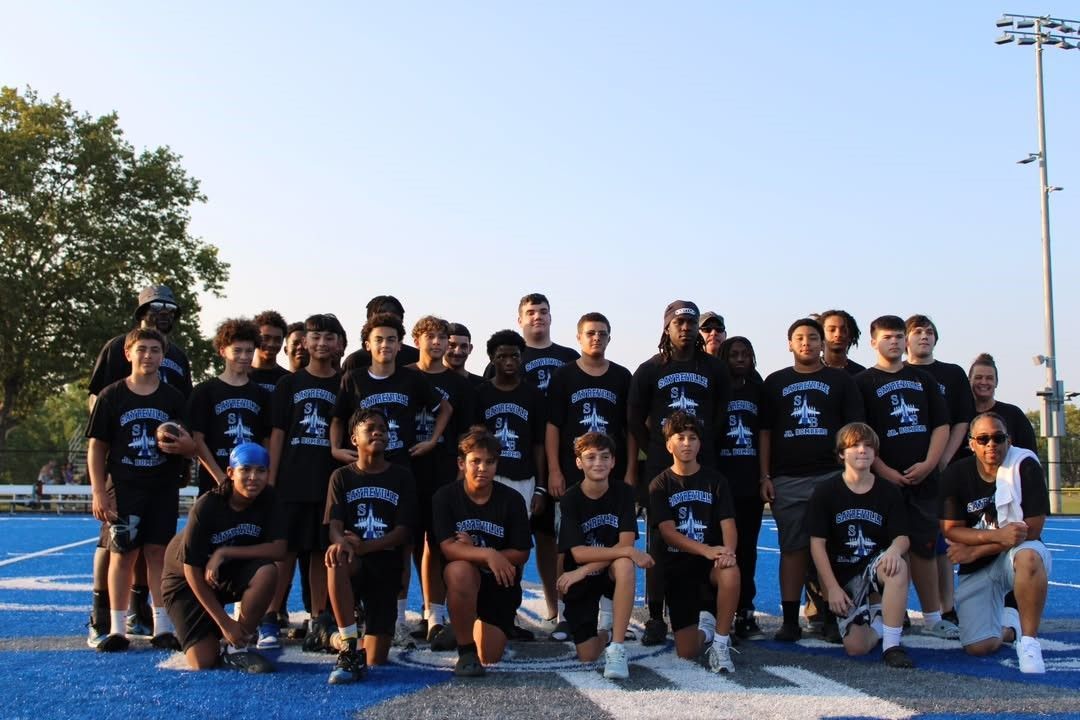 Football team poses on a blue field. Players wear black shirts with logo. Sunny, outdoor setting.