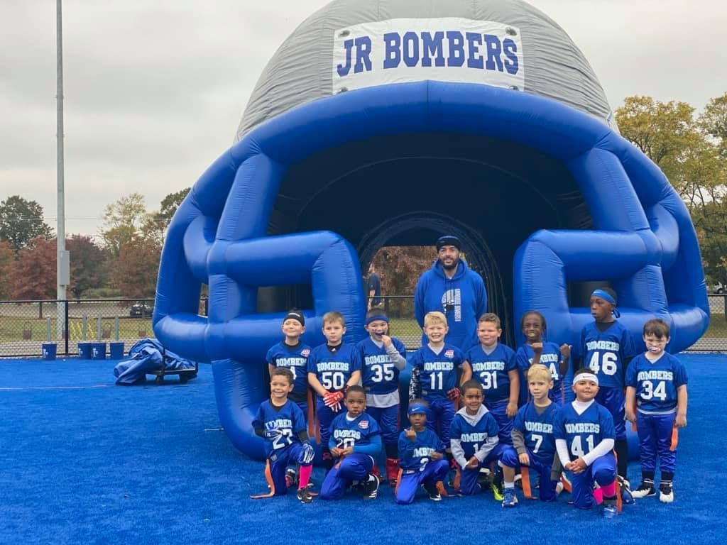Youth football team, the Jr. Bombers, pose in front of a blue helmet tunnel on a blue field.