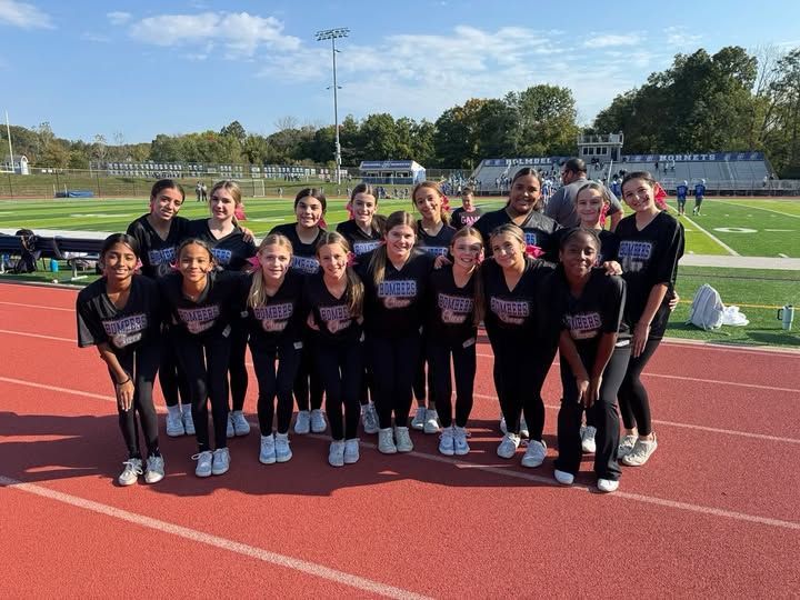 A group of cheerleaders in black uniforms poses on a track at a football field, smiling on a sunny day.