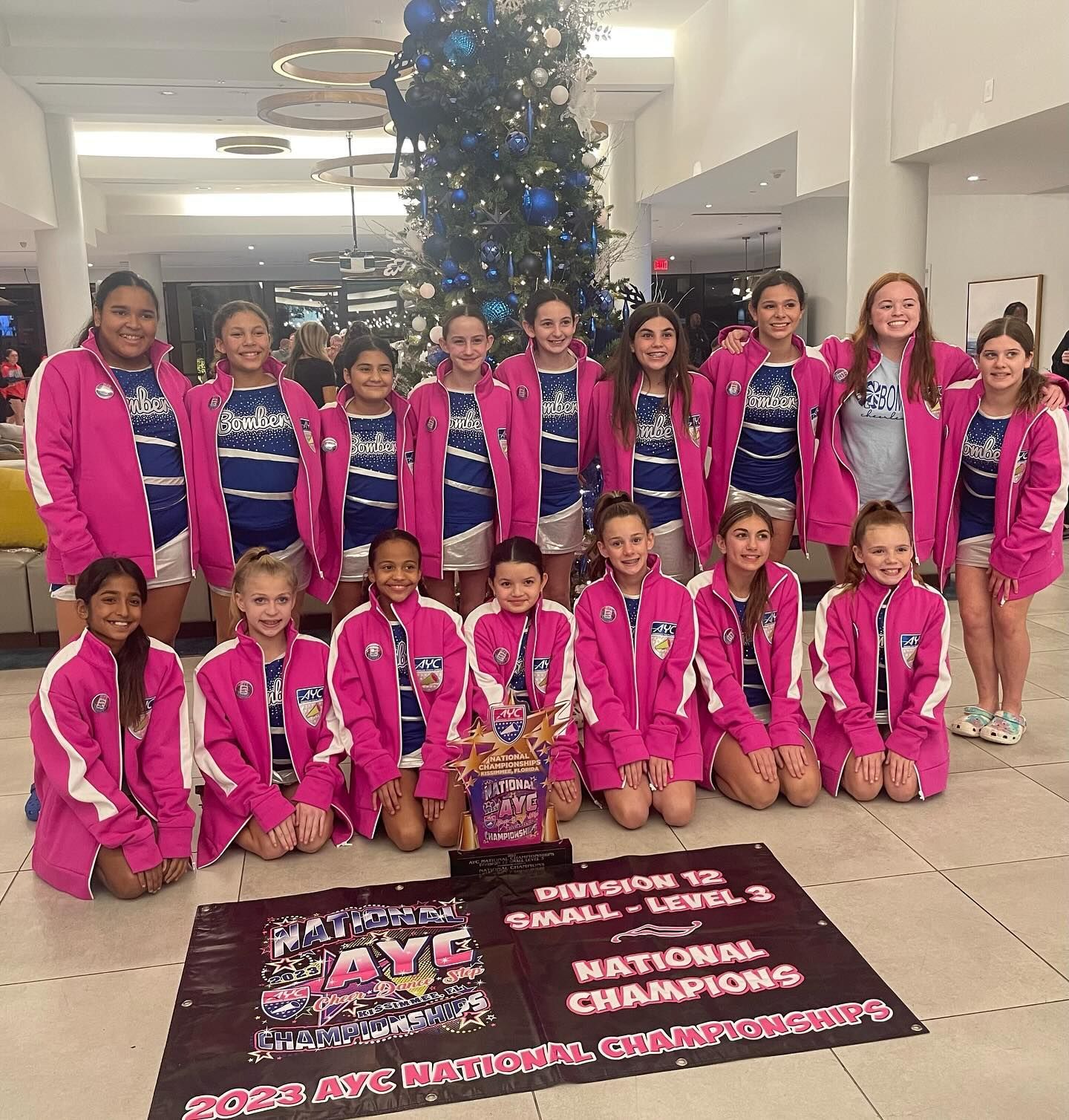 Cheerleading team in pink jackets, posing with trophy and banner at event.