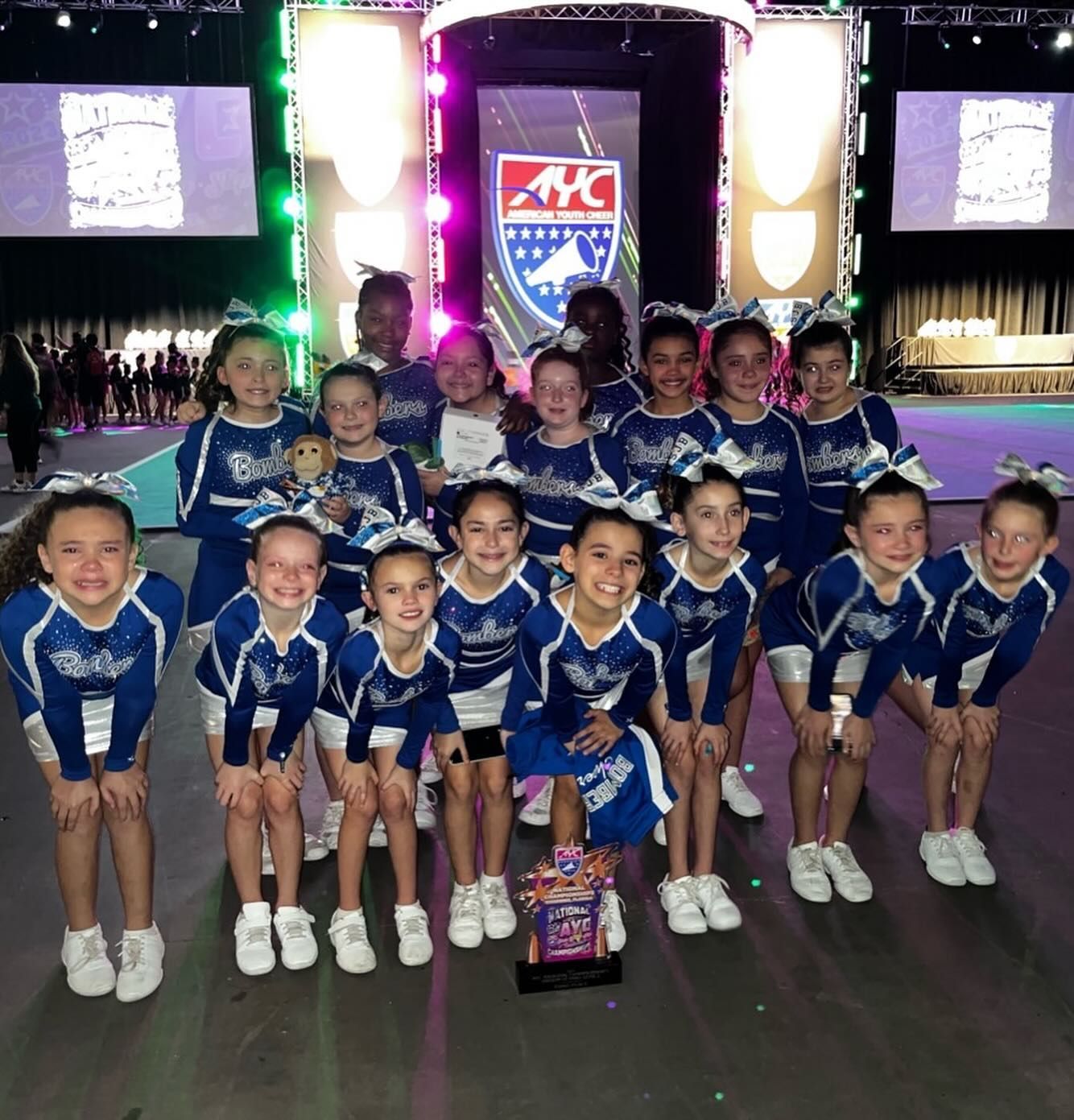 Youth cheerleading team in blue uniforms poses with a trophy.