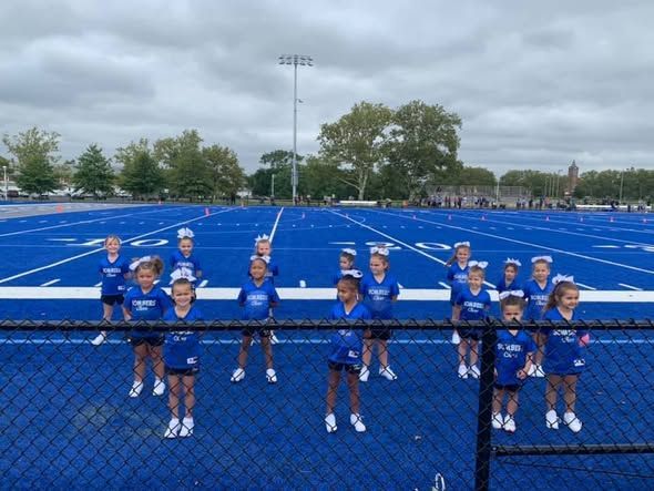 Cheerleaders in blue uniforms on a blue football field with white yard lines, overcast sky.