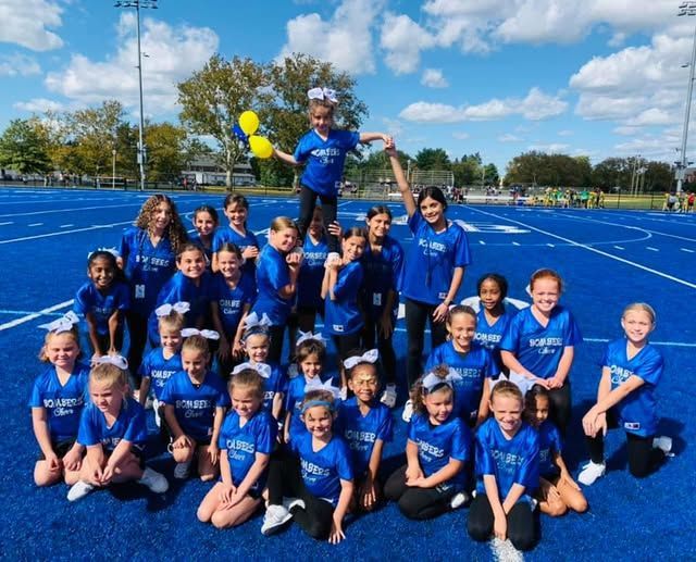 Youth cheerleaders in blue uniforms on a blue track, some kneeling, one on top holding pom poms.