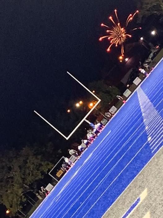 Football field at night with fireworks over the goalposts. Blue track, crowd, and dark sky.