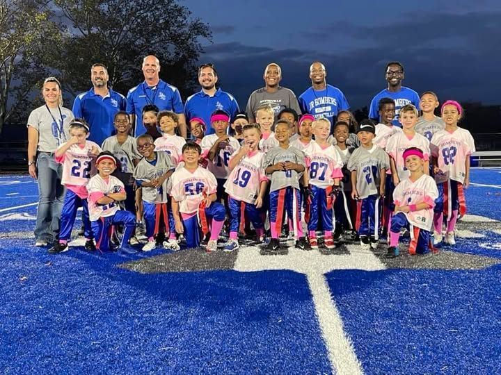 Youth flag football team posing on a blue field with coaches at night. Players wear pink and blue.