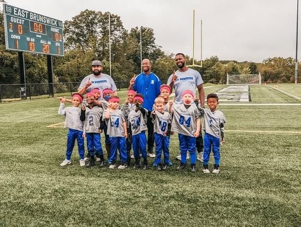 Youth football team posing on a field with coaches and scoreboard. Players wearing blue and gray.