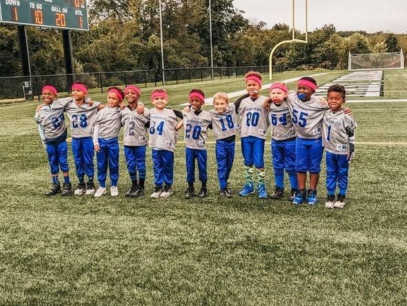 Youth football team in blue and grey uniforms on a green field. Players huddle, arms around each other.
