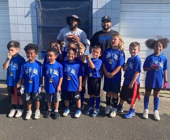 Youth flag football team in blue jerseys poses with coaches outside.