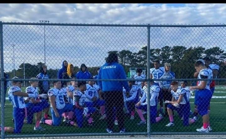 Football team kneeling, praying on a green field. Players wearing blue and white uniforms, coach in blue jacket.