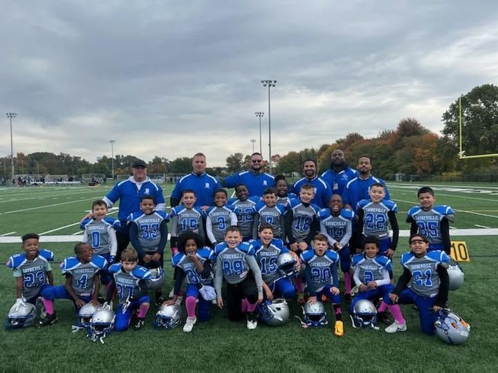 Youth football team posing on a green field. Players in blue and gray uniforms. Coaches stand behind them.