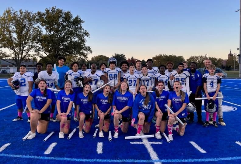 Football team and cheerleaders pose on a blue field, holding brooms.