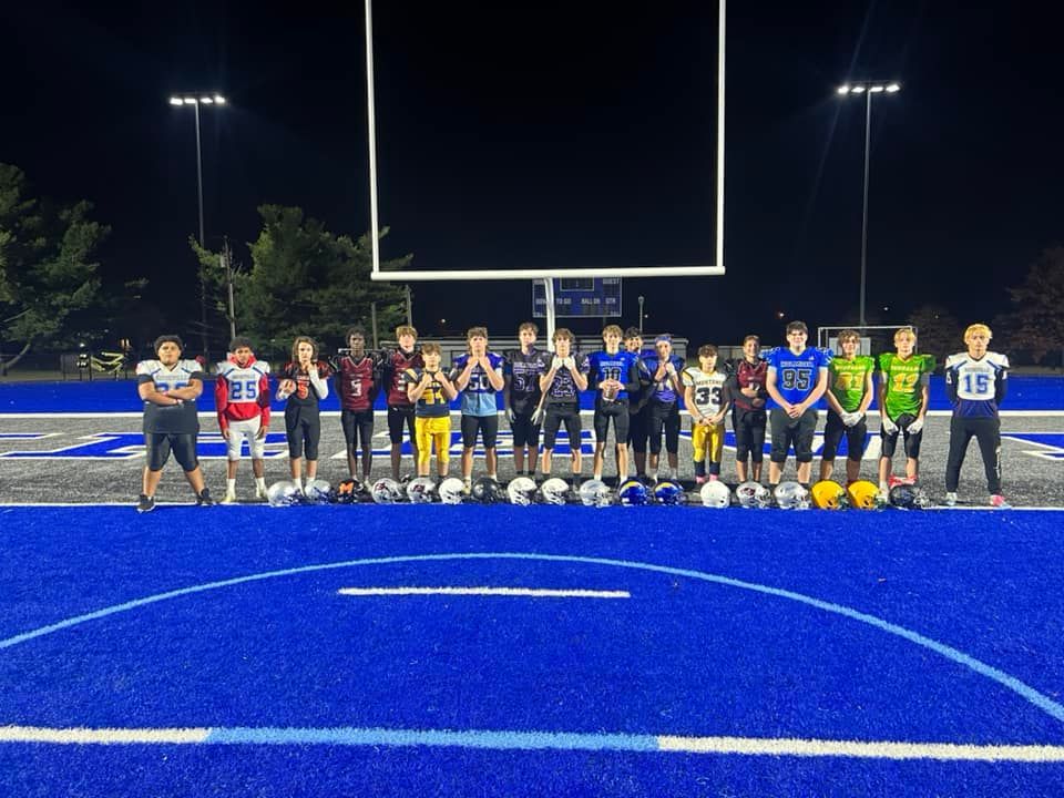 Football team stands on a blue field at night in front of the goal post. Helmets are lined up in front of the players.
