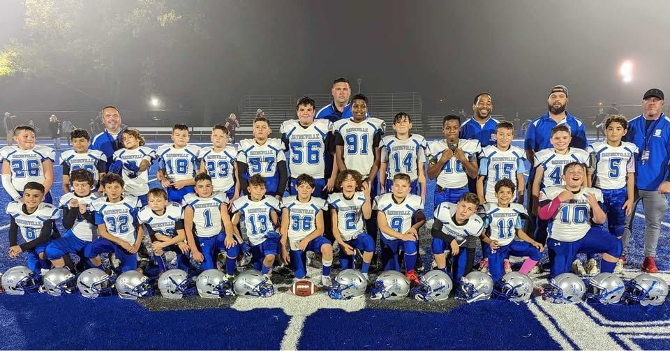 Youth football team in blue and white uniforms, posing with coaches on a blue field at night.